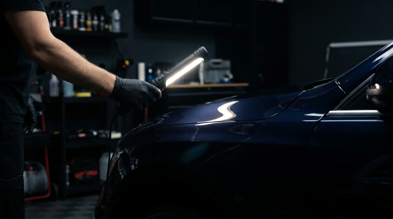 A gloved hand running a hard LED inspection light across a freshly coated dark navy fender in the Lustre bay