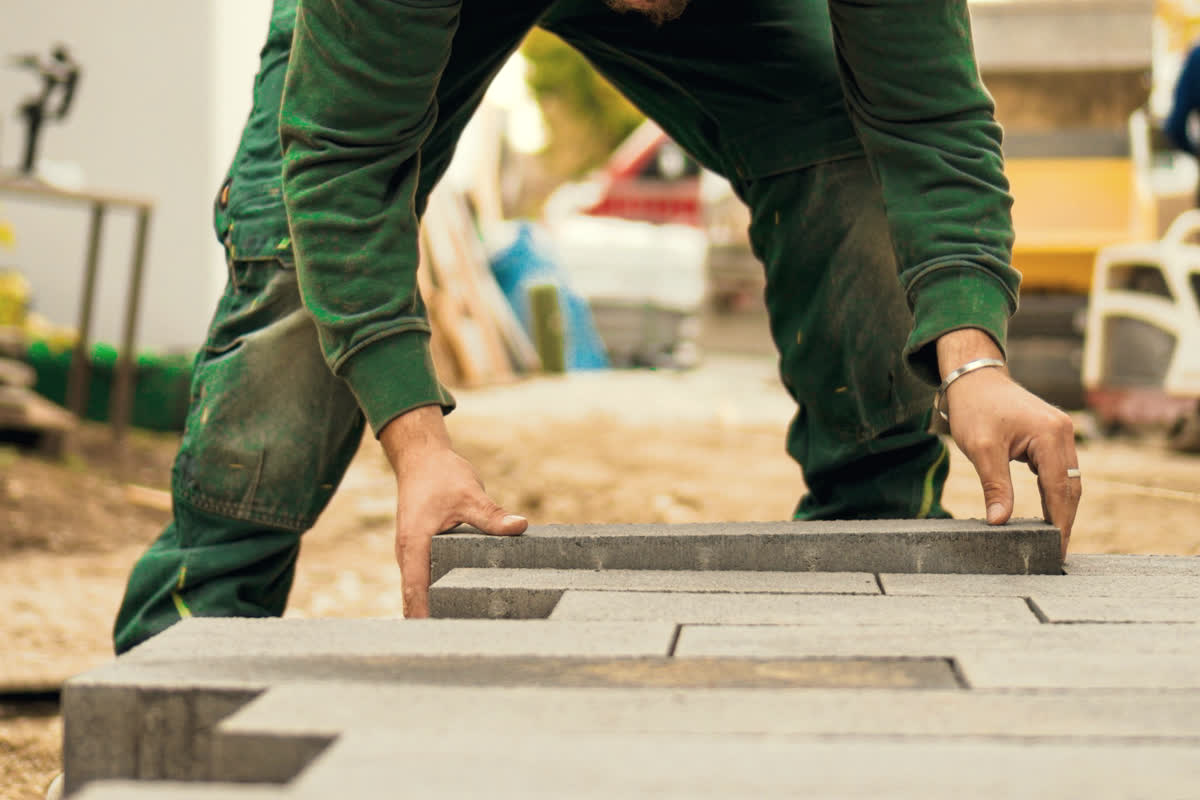 A worker in green work trousers, bent forward, lowering a long grey concrete paving slab onto a bedding layer beside an already-laid course.