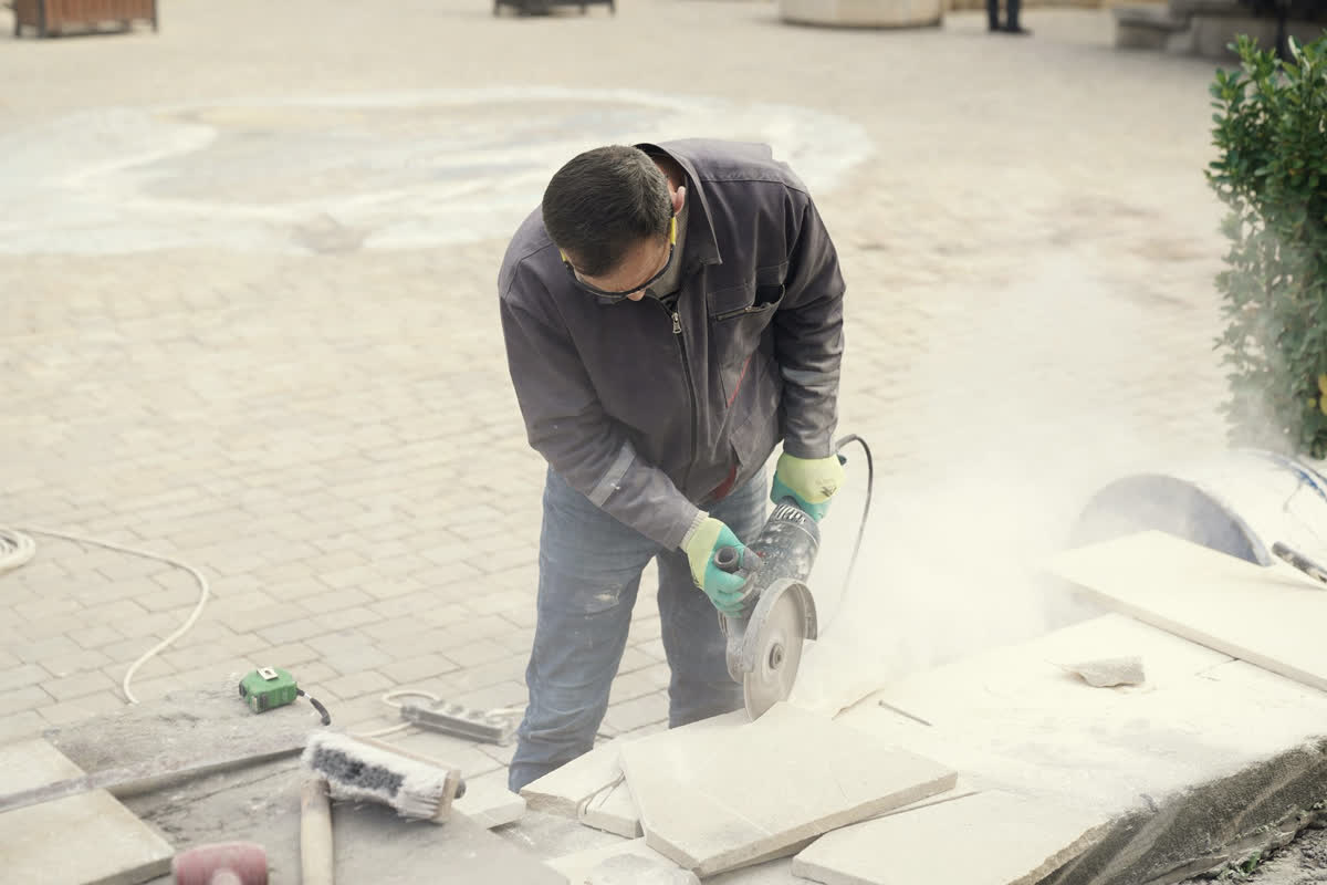 A worker in a dust-covered jacket, safety glasses and ear plugs, making a cut with a handheld saw, dust hanging in the air.