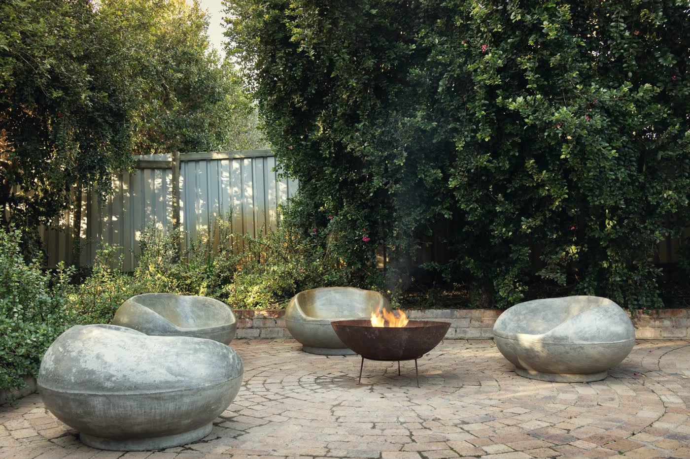 A circular tumbled-cobble patio laid in a radial fan pattern, with a lit steel fire bowl at its centre and four cast-concrete boulder-form chairs around it.