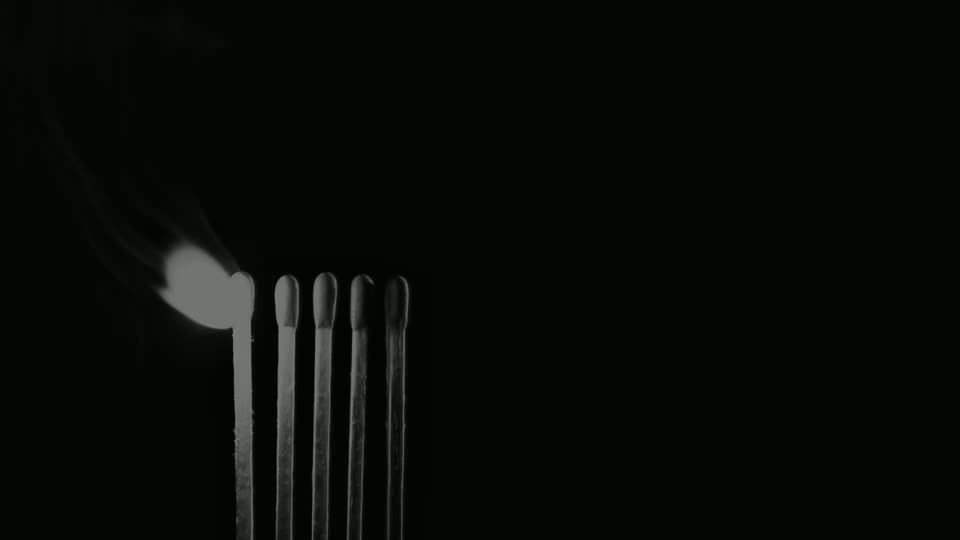 Frame 001 of the match-strike sequence: five match heads standing in a row, none of them alight. A soft grey glow sits on the leftmost head and a single thread of smoke curls up and away to the left. The right two-thirds of the frame is empty black.
